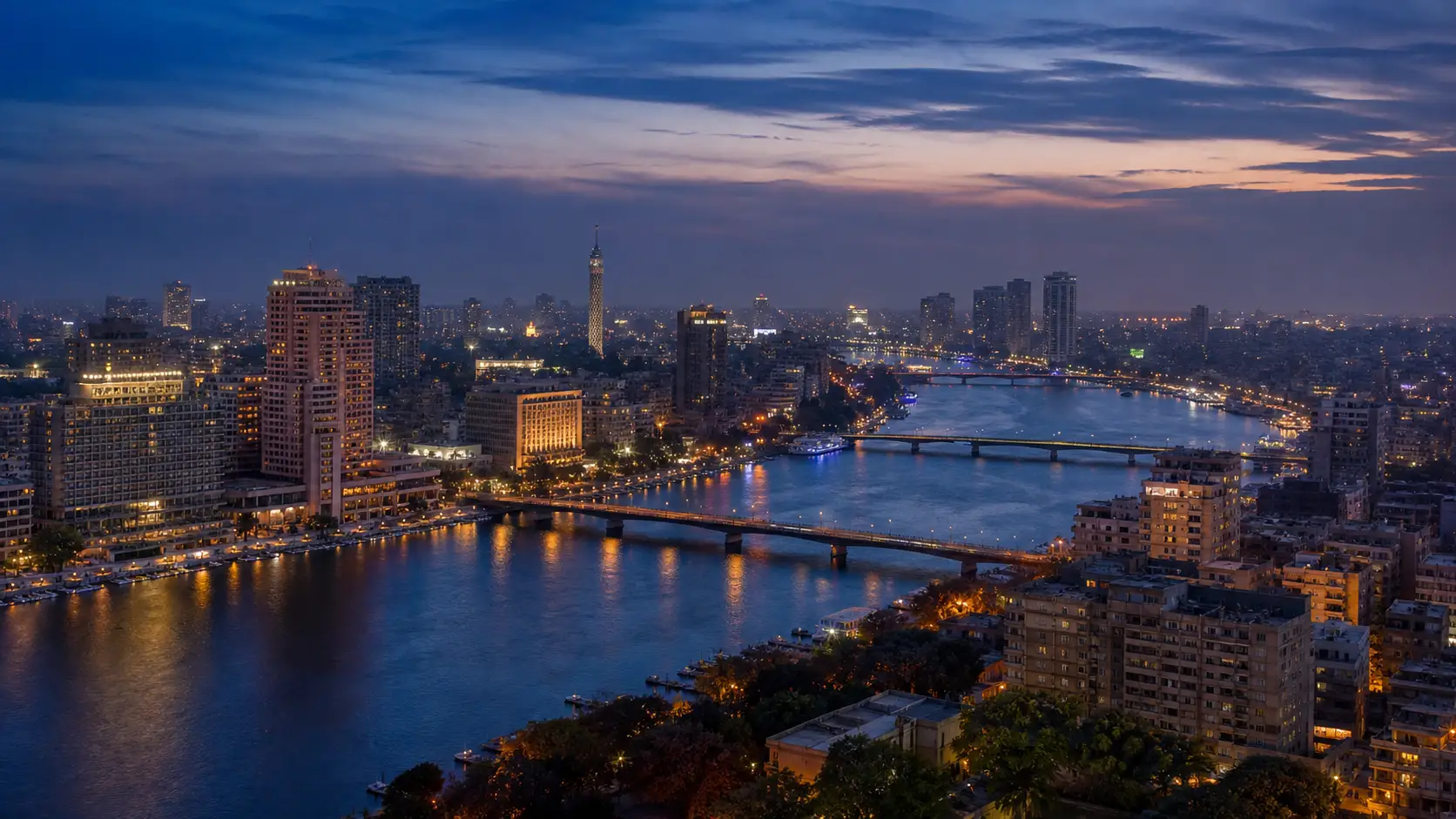 The Nile curves through the illuminated Cairo skyline at blue hour, with bridges and the Cairo Tower visible beneath a darkening sky.